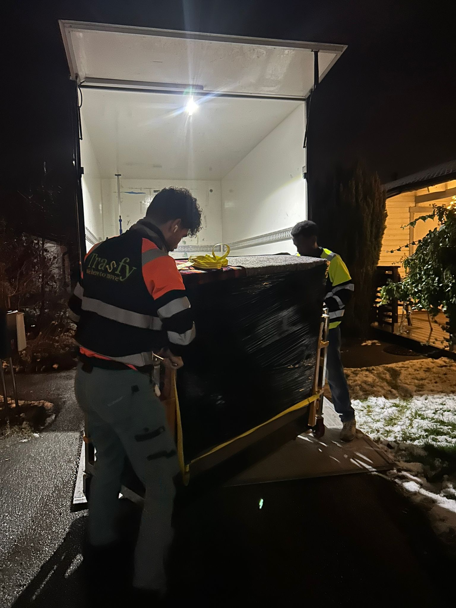 Flyttare rullar ett inplastat piano mot flyttbilen en vinterkväll