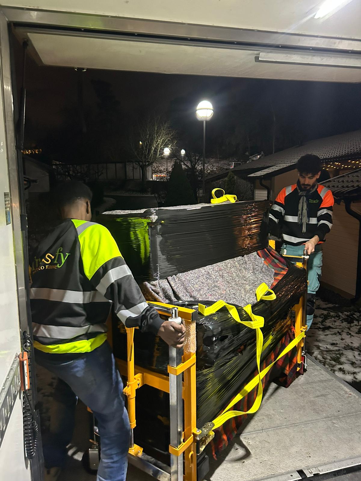 Två flyttare i varselväst säkrar ett piano på pianovagn vid lastbilen